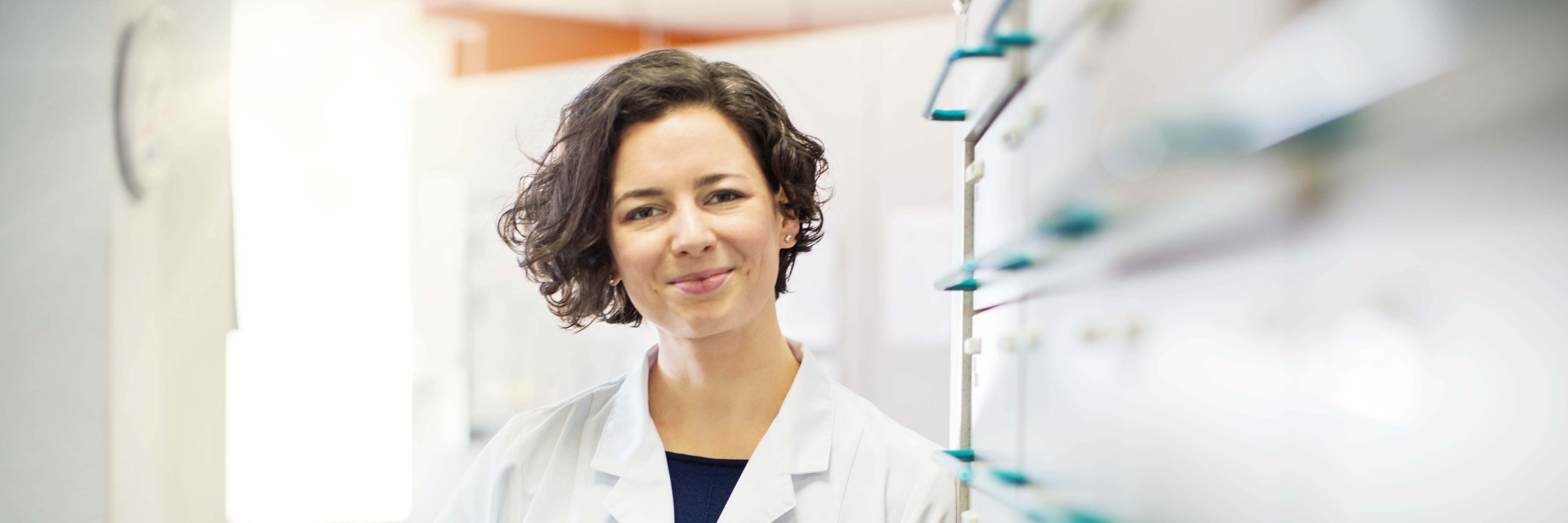 Middle-aged female clinician in a white lab coat, standing in a medical office next to some filing cabinets